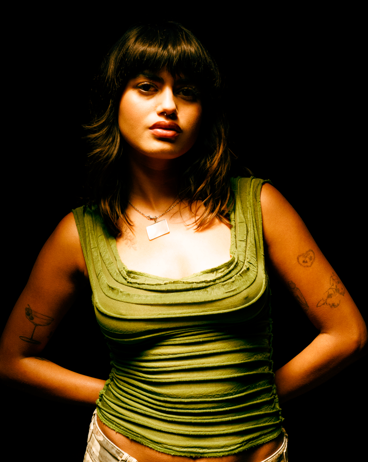 Young woman with dark hair and bangs, wearing a green tank top with layered fabric details, standing against a black background.