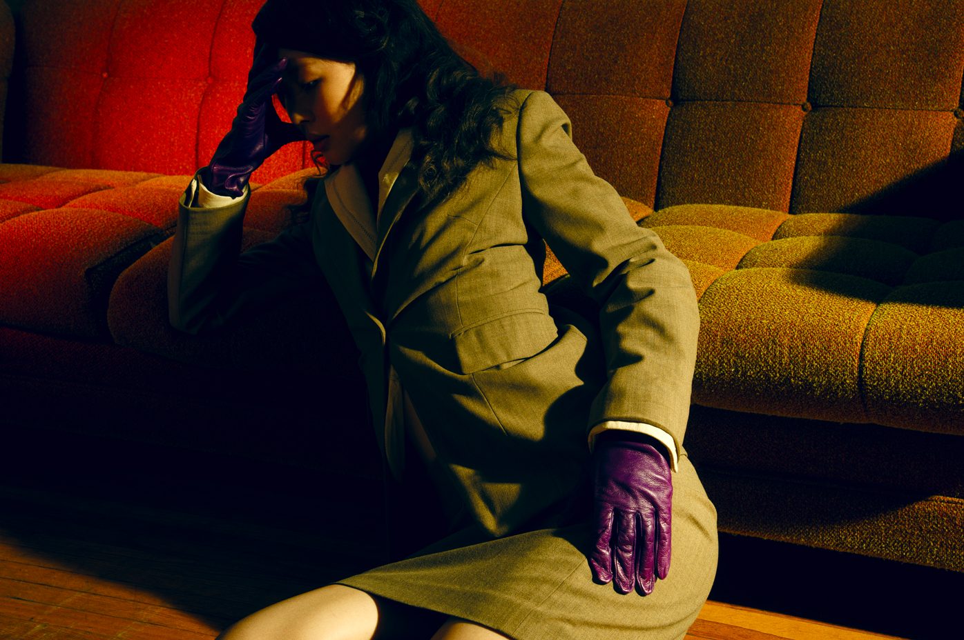 A woman in a beige suit with purple gloves sitting on the floor leaning against a vintage red and orange patterned sofa, holding her head with her left hand.