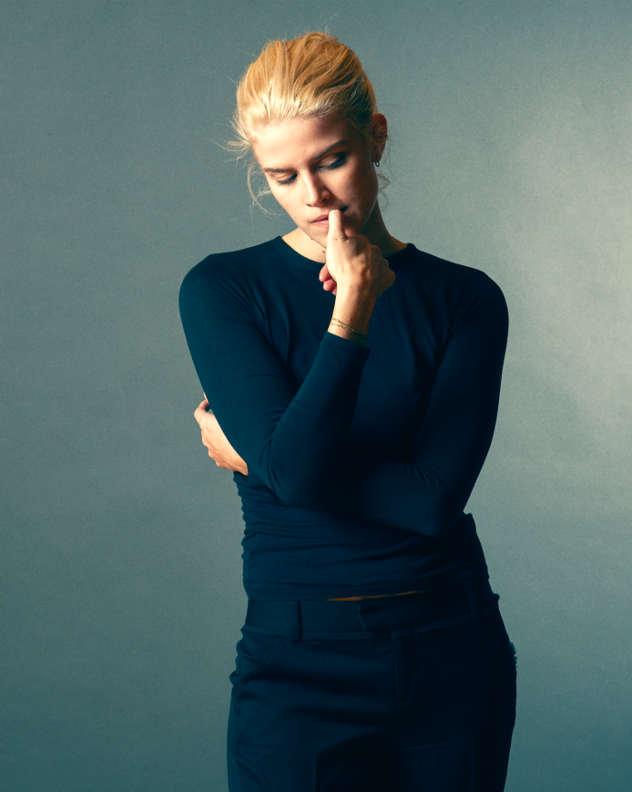 A young woman with blonde hair, wearing a dark long-sleeve shirt and pants, is standing with her eyes closed, touching her lips with her finger, and her arms crossed in front of her body against a plain gray background.