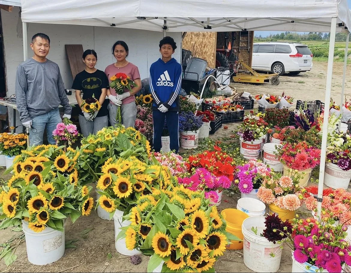 Four people standing behind a stall filled with colorful flowers including sunflowers and pink blooms, under a white canopy outdoors near a white van and a construction area.