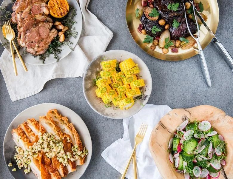 Assorted dishes on a dining table, including roasted meat with vegetables, a salad with radishes and greens, a bowl of sliced yellow zucchini, a breaded chicken cutlet with white sauce and herbs, and another salad with greens and radishes.