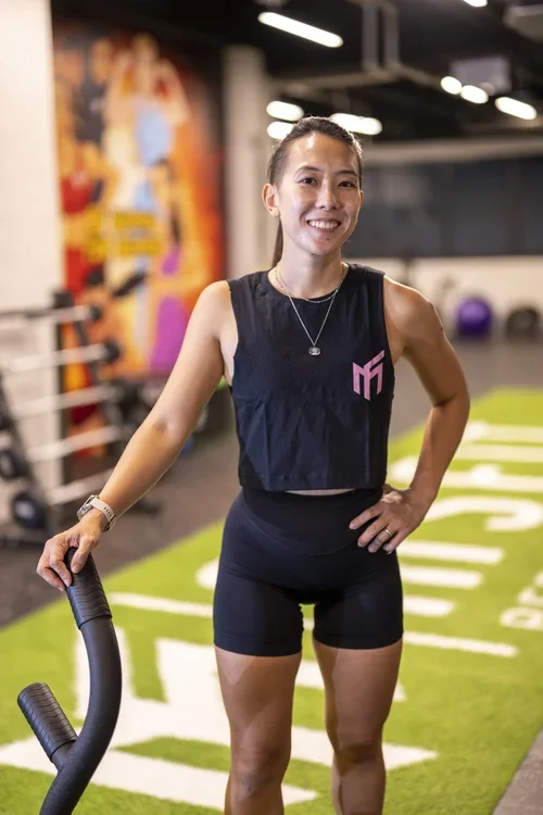 A woman in workout clothes standing in a gym, smiling, with her hand on a piece of exercise equipment.