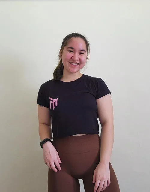 A young woman wearing a black t-shirt and brown pants, standing against a plain off-white wall, smiling at the camera.