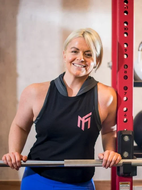 A woman smiling in a gym, holding a barbell, wearing a black athletic tank top with a pink logo and blue workout pants.