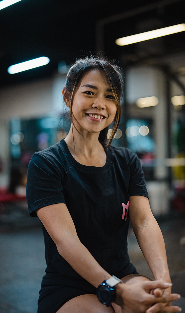 Young woman with dark hair tied back, smiling, wearing a black t-shirt and smartwatch, in a gym or fitness center with exercise equipment and soft lighting.