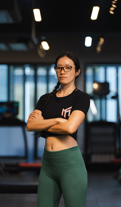 A woman with glasses, wearing a black crop top and green workout leggings, standing with arms crossed in a gym.