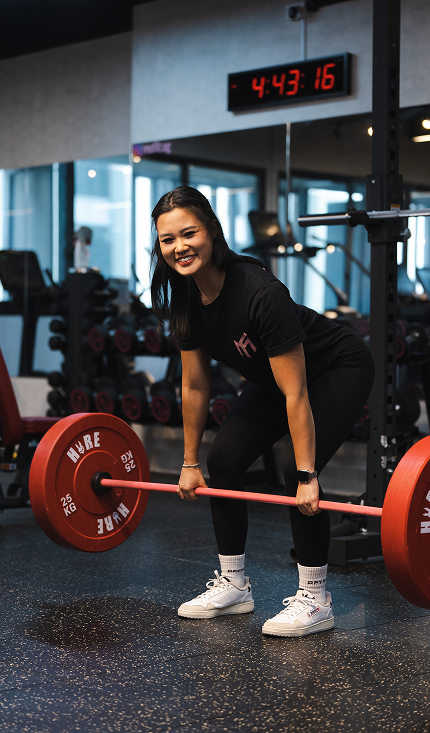 A young woman lifting a barbell with red weight plates in a gym, smiling at the camera.