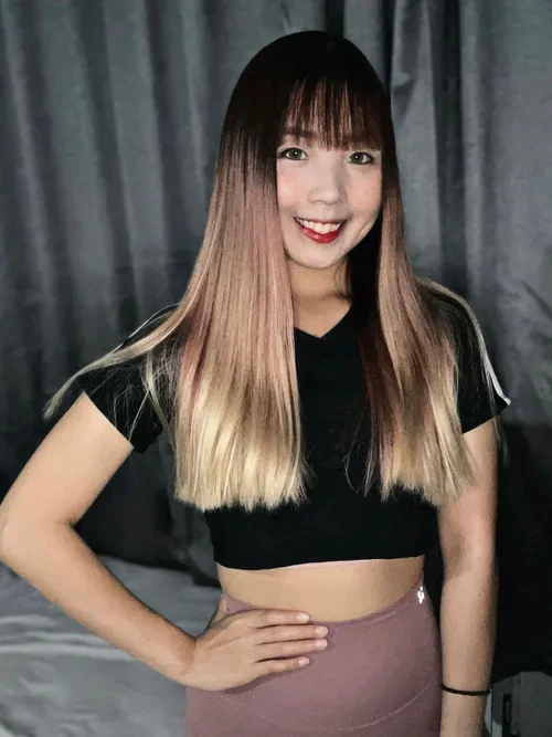 A young woman with long, ombre hair from dark brown to blonde, smiling and posing indoors in front of a dark curtain, wearing a black cropped T-shirt and mauve high-waisted pants.