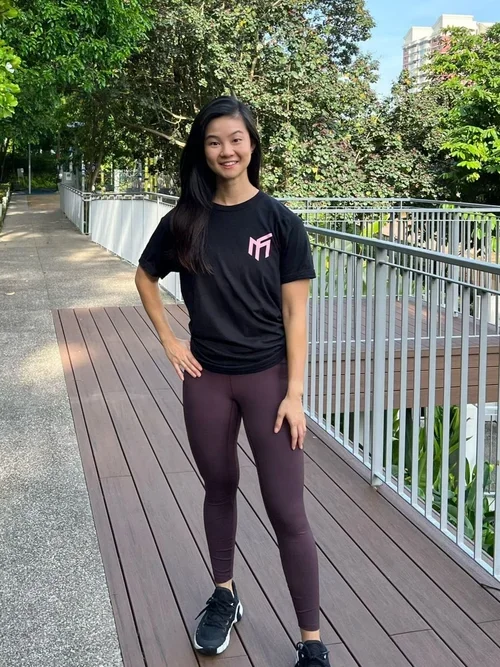 Young woman in athletic outfit standing on wooden pathway outdoors, smiling, with trees and buildings in the background.