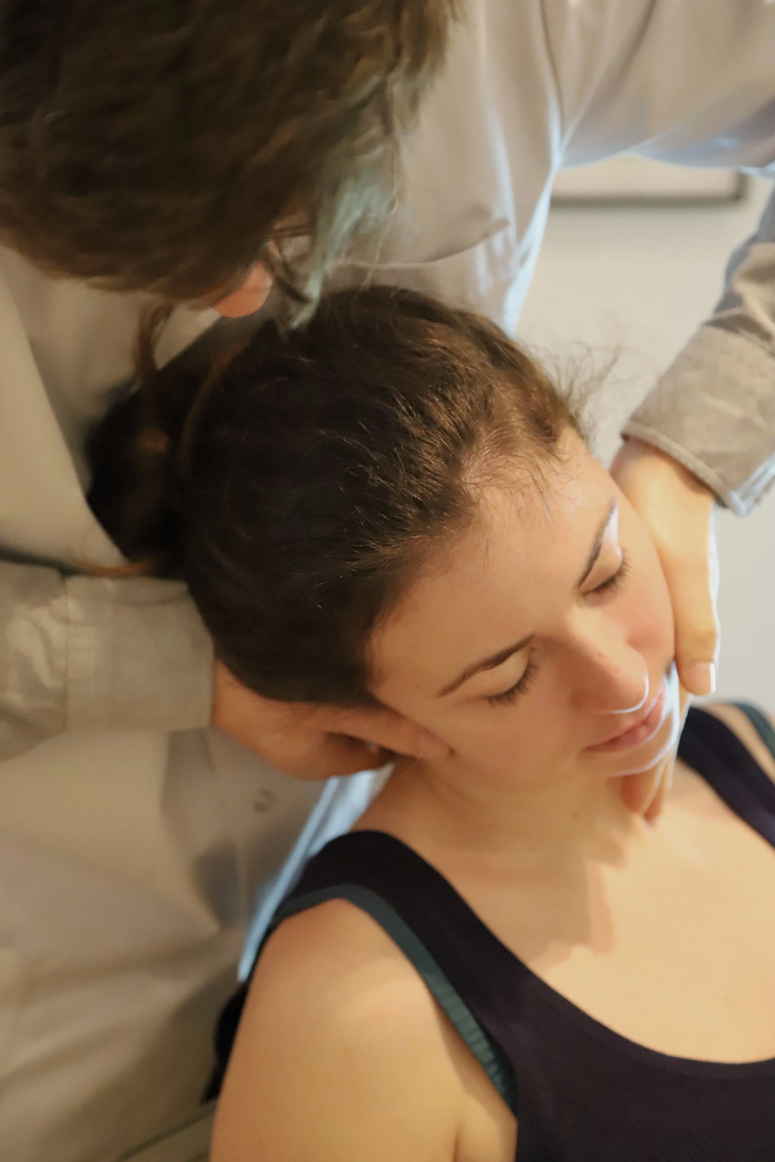 Deux femmes en coiffure épaulent une jeune femme, qui sourit doucement, lors d'un massage ou séance de relaxation, dans un cadre intérieur.