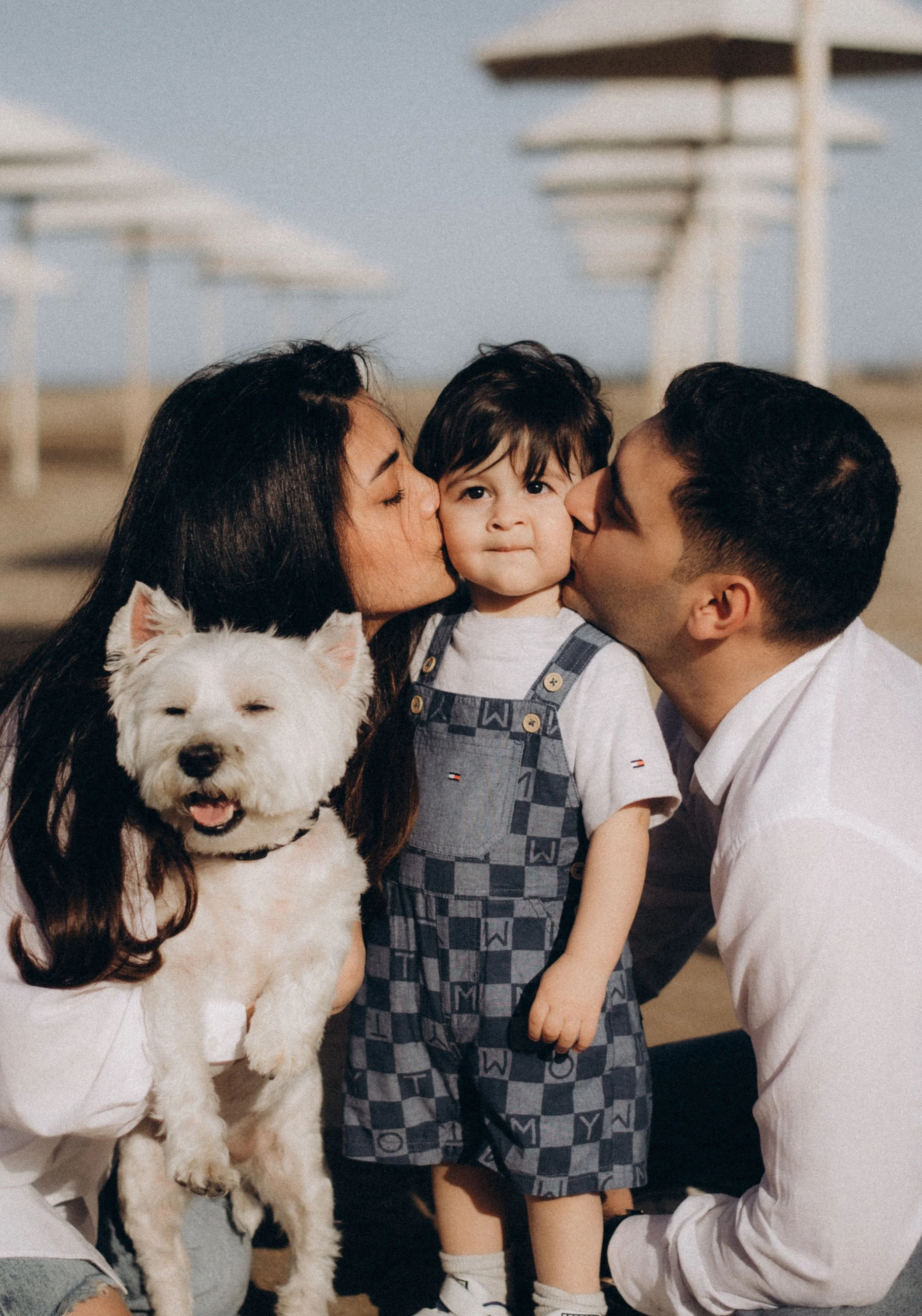 A mother and father kissing their toddler on the cheeks while the mother holds their small white dog
