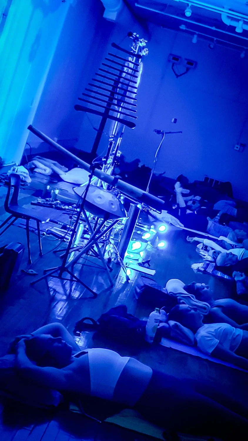 A large group setting showing various musical instruments on stands and the floor, illuminated by blue lights, with people lying down or resting on the floor experiencing a sound journey with gongs, didgeridoo, handpan and more.