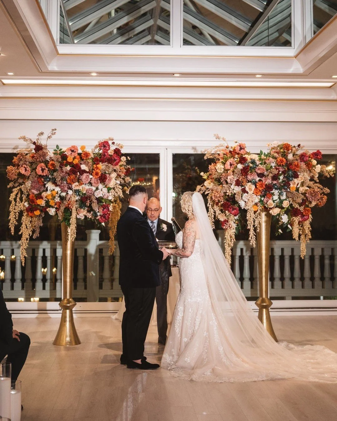 G + E's wedding ceremony and reception florals spoke of quiet luxury: lush florals, a rich color palette, and shining gold accents.

They exchanged vows beneath two lush floral installations overflowing with rich seasonal color. Deep reds, burnt oran