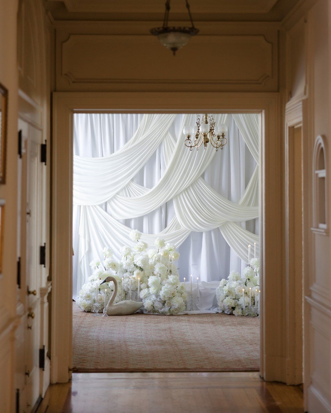 A quiet reveal into something timeless: layers of soft draping cascading like silk, a stage framed in delicate whites, and the bride pictured among blooms as if pulled from a dream. Fluffy white hydrangea, crisp white roses, and luxe white peonies de