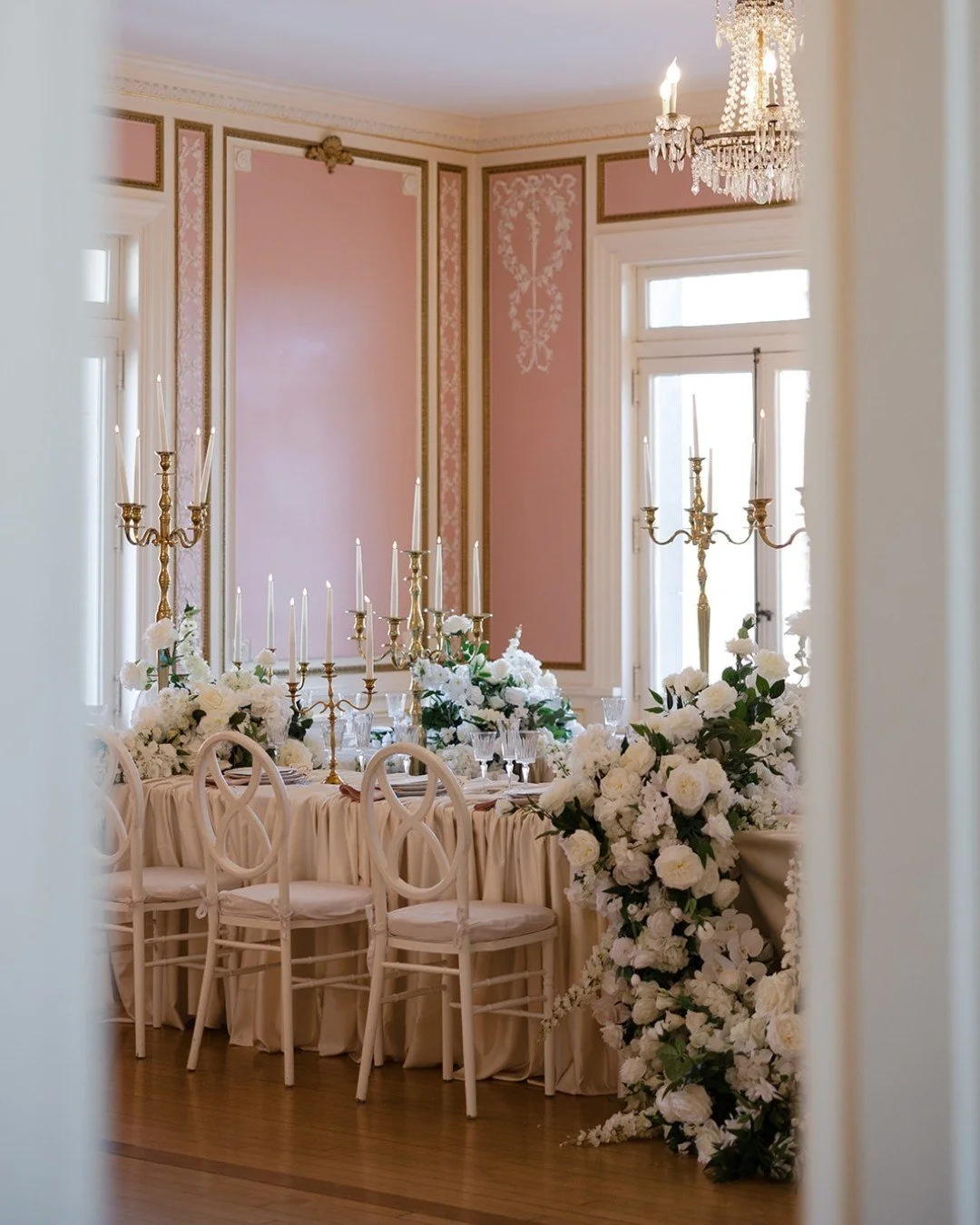 Blush walls, warm golden candelabras glowing softly, and layers of lush white florals weaving their way down a beautifully draped table...every detail designed to feel timeless, romantic, and effortlessly refined.

Crystal glassware catches the candl
