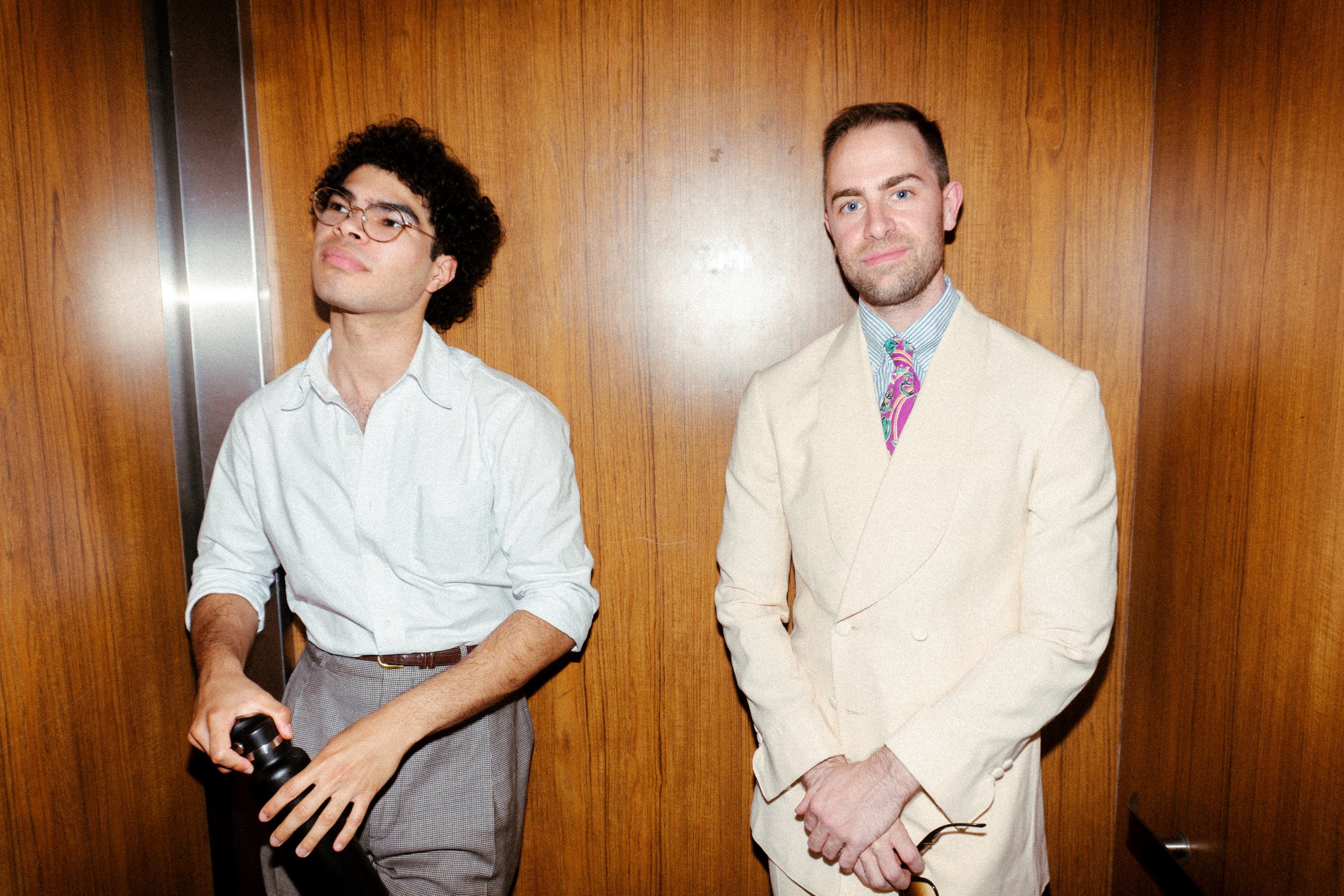 Two men in an elevator with a wooden wall background, one wearing a white shirt and glasses holding a bottle, the other in a beige suit with a colorful tie.