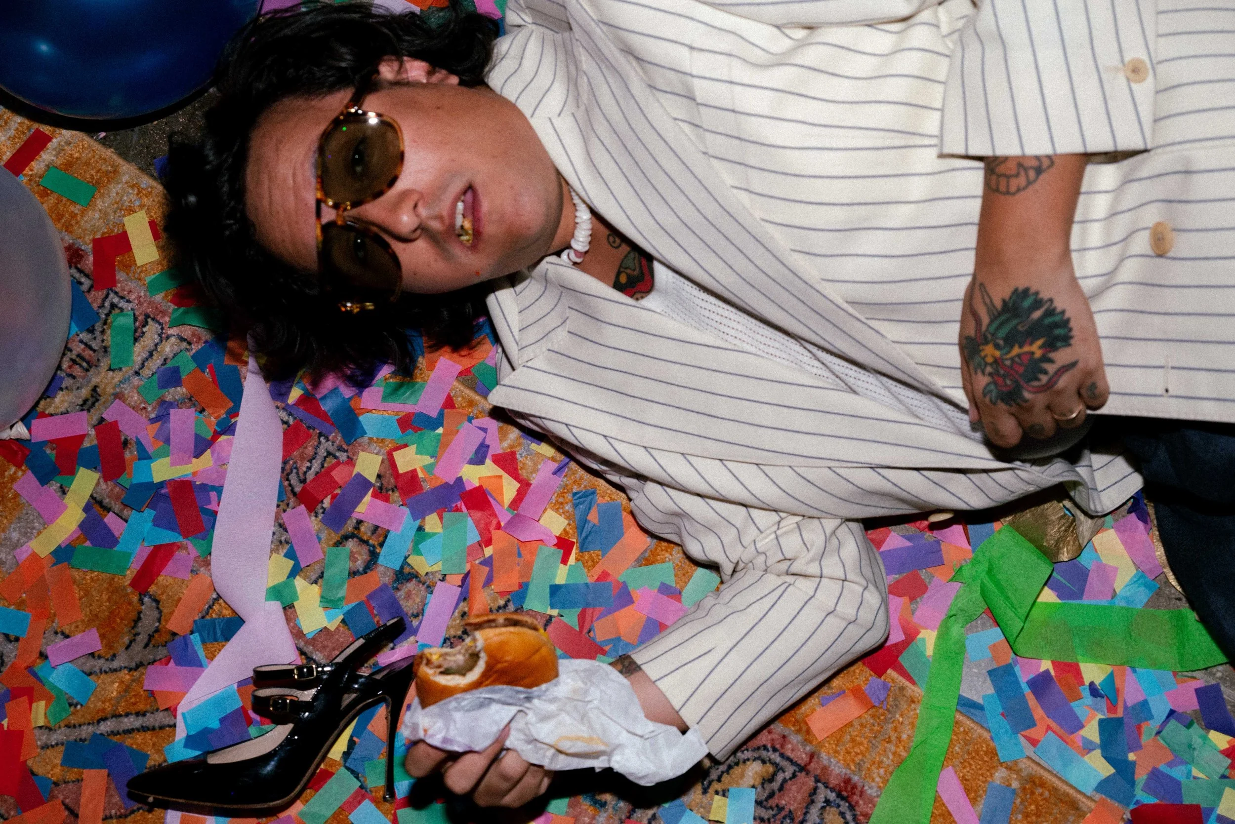 A man with dark curly hair, wearing sunglasses, a white jacket with pinstripes, and jewelry, is lying on a colorful, confetti-covered floor. He is holding a partially eaten sandwich in one hand and is surrounded by multicolored paper strips and ballo