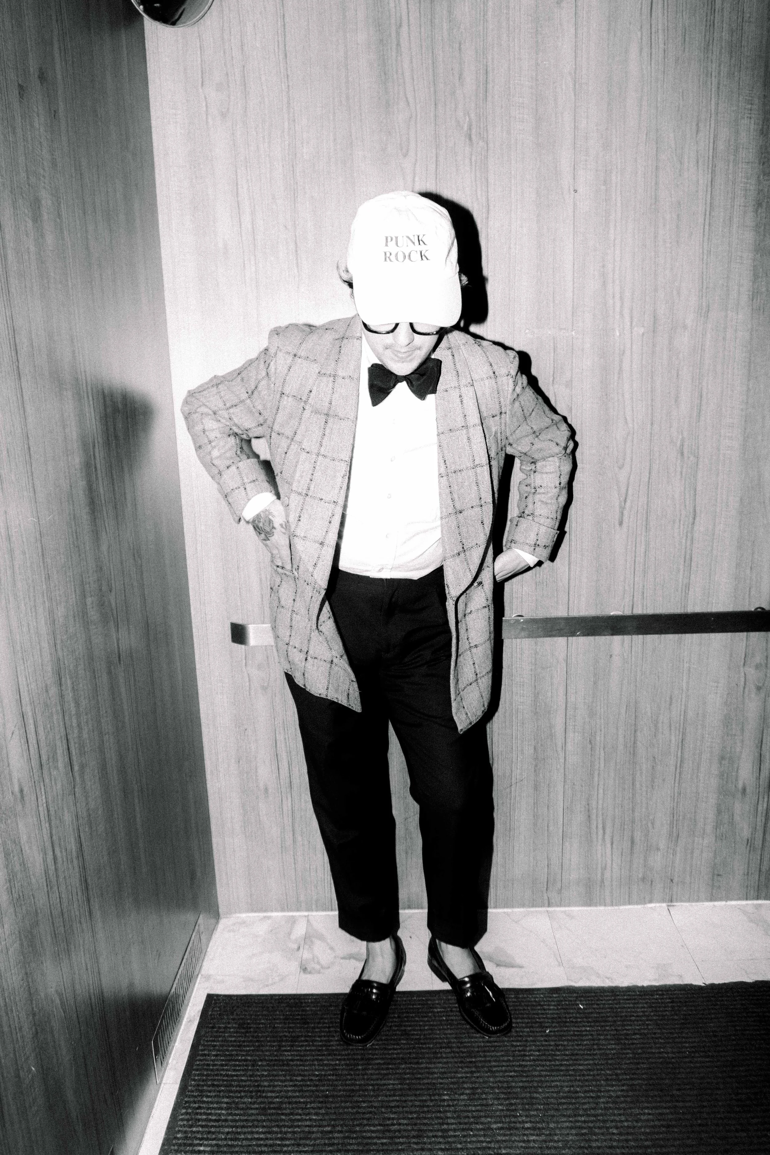 A man wearing a baseball cap that says 'PUNK ROCK', glasses, a bowtie, a checkered blazer, white shirt, black pants, and loafers stands with his head tilted down and hands in his pockets in an elevator with wooden paneling.