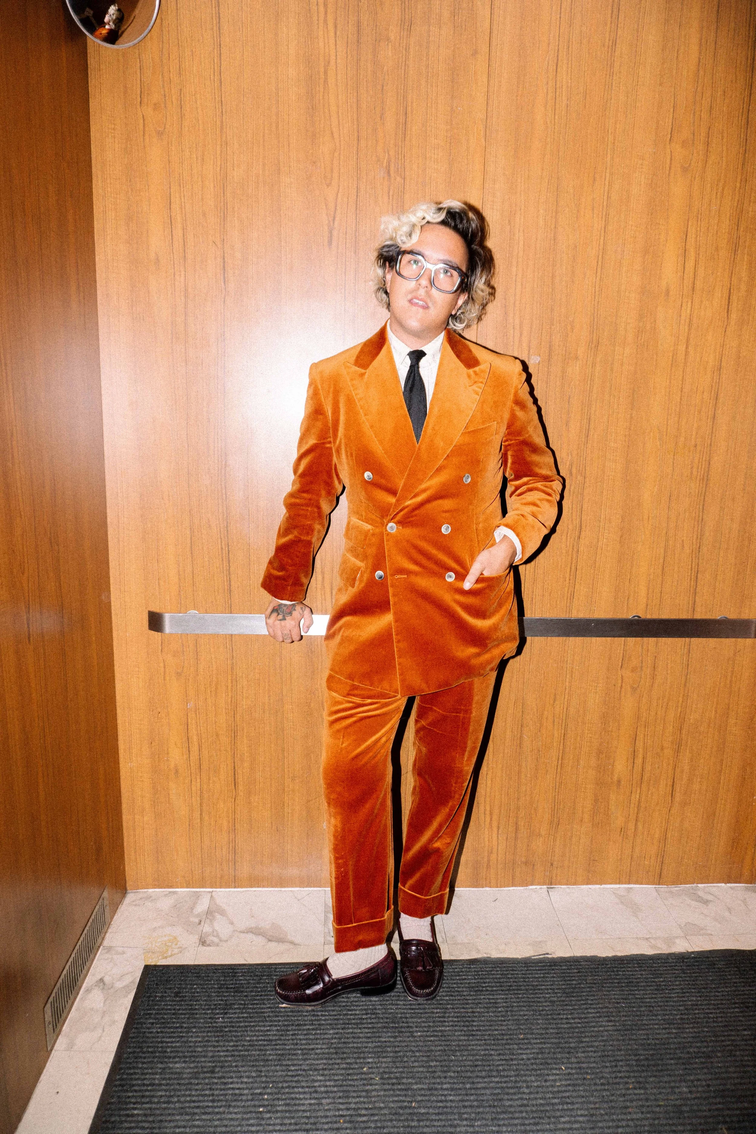 A man in an orange velvet double-breasted suit, white shirt, black tie, and glasses standing in front of a wooden wall, leaning against an elevator door.