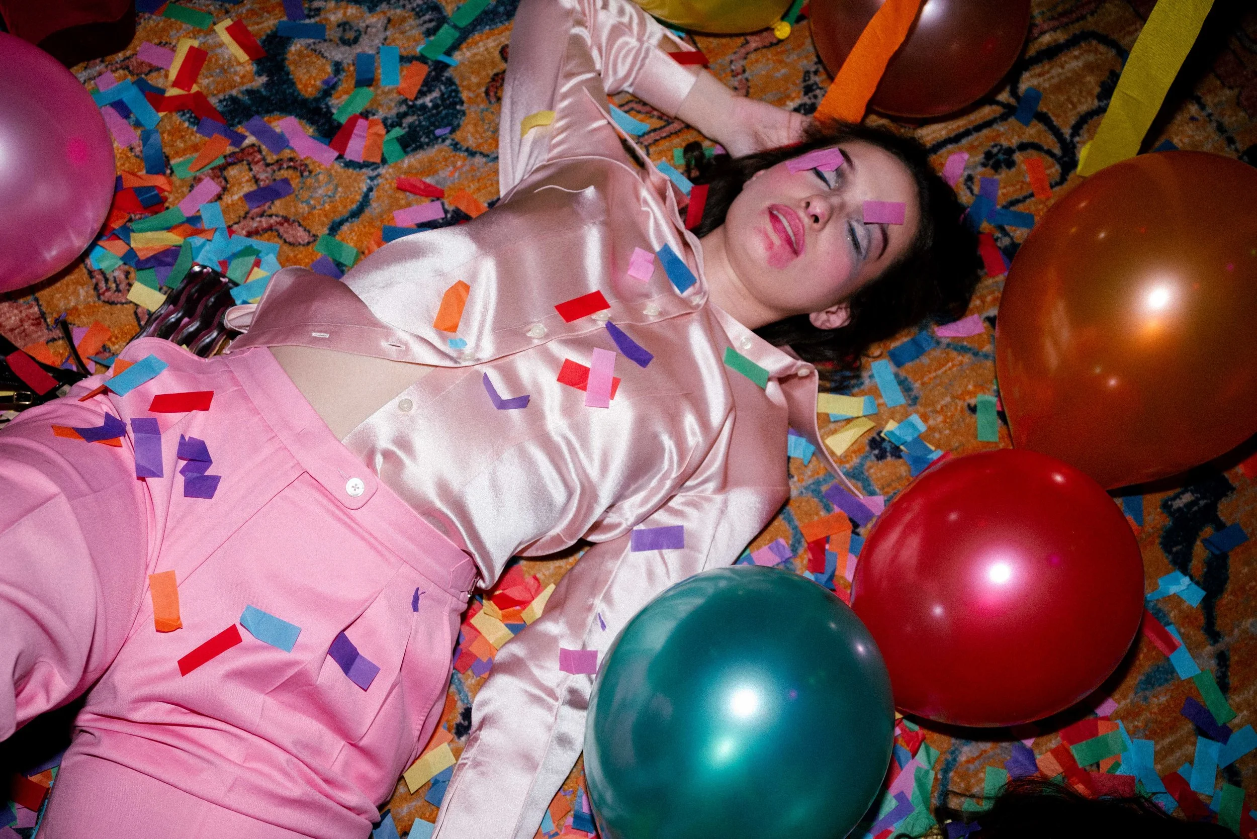 A woman in a pink satin outfit lying on a colorful carpet surrounded by balloons and colorful confetti with her eyes closed.