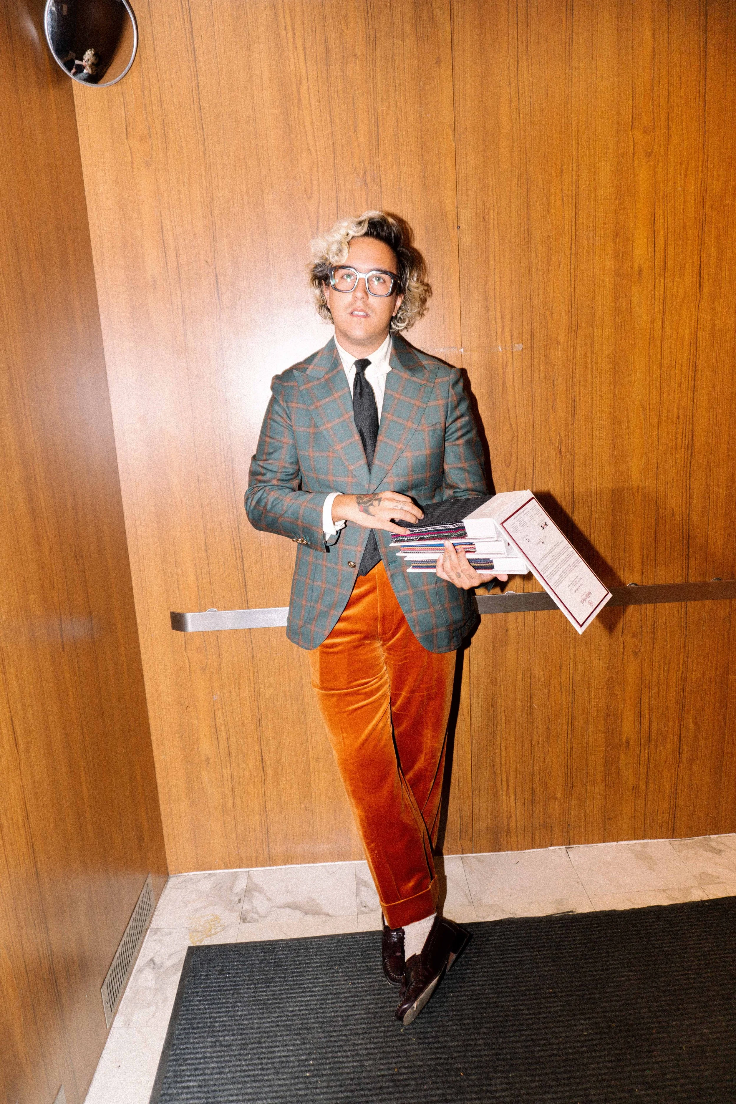 A person standing in an elevator holding a stack of papers and folders, wearing glasses, a plaid blazer, orange velvet pants, and proof shoes, against a wood-paneled wall.