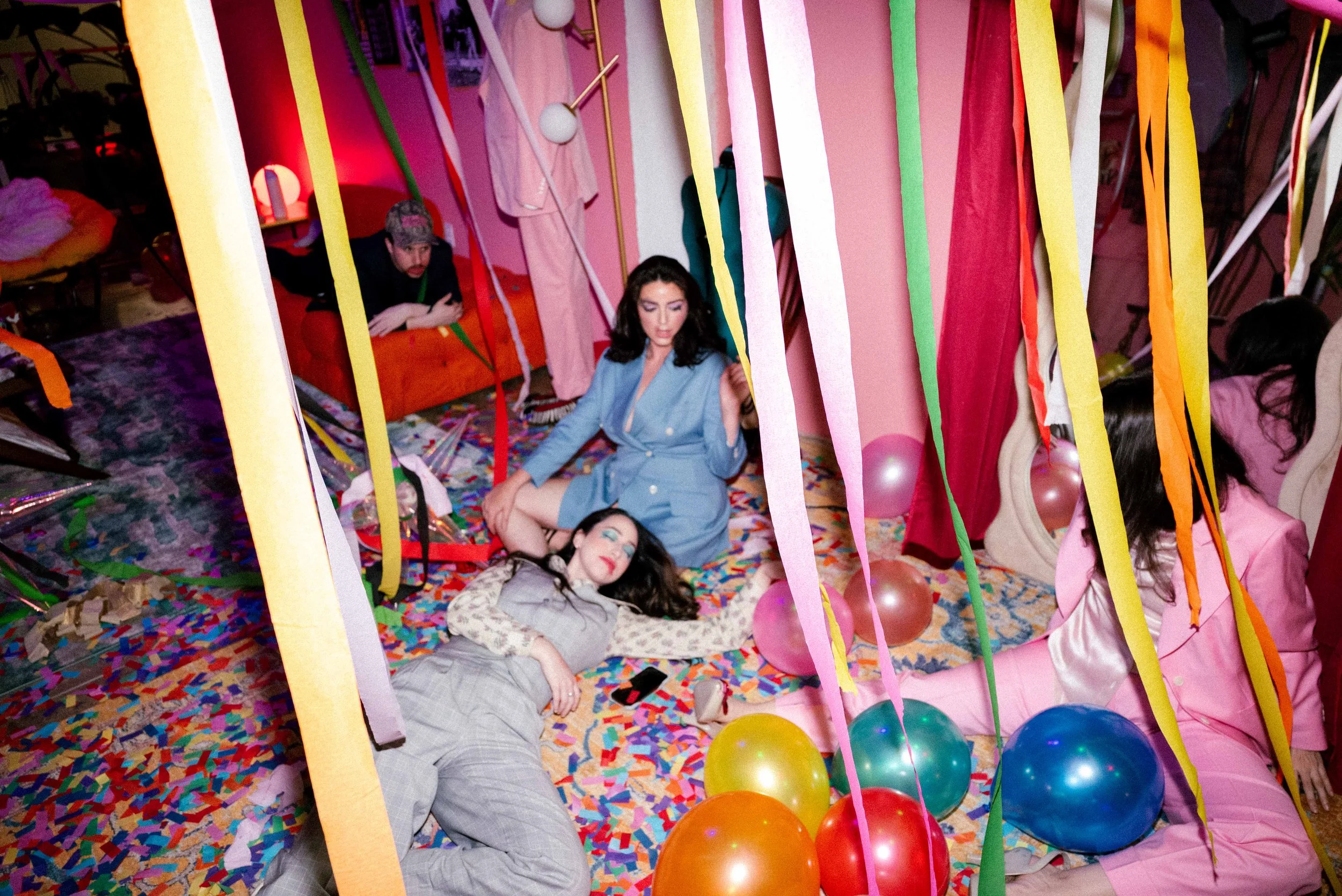 Group of people at a colorful party with balloons, streamers, and confetti. Some are sitting, some lying down, in a room decorated for celebration.