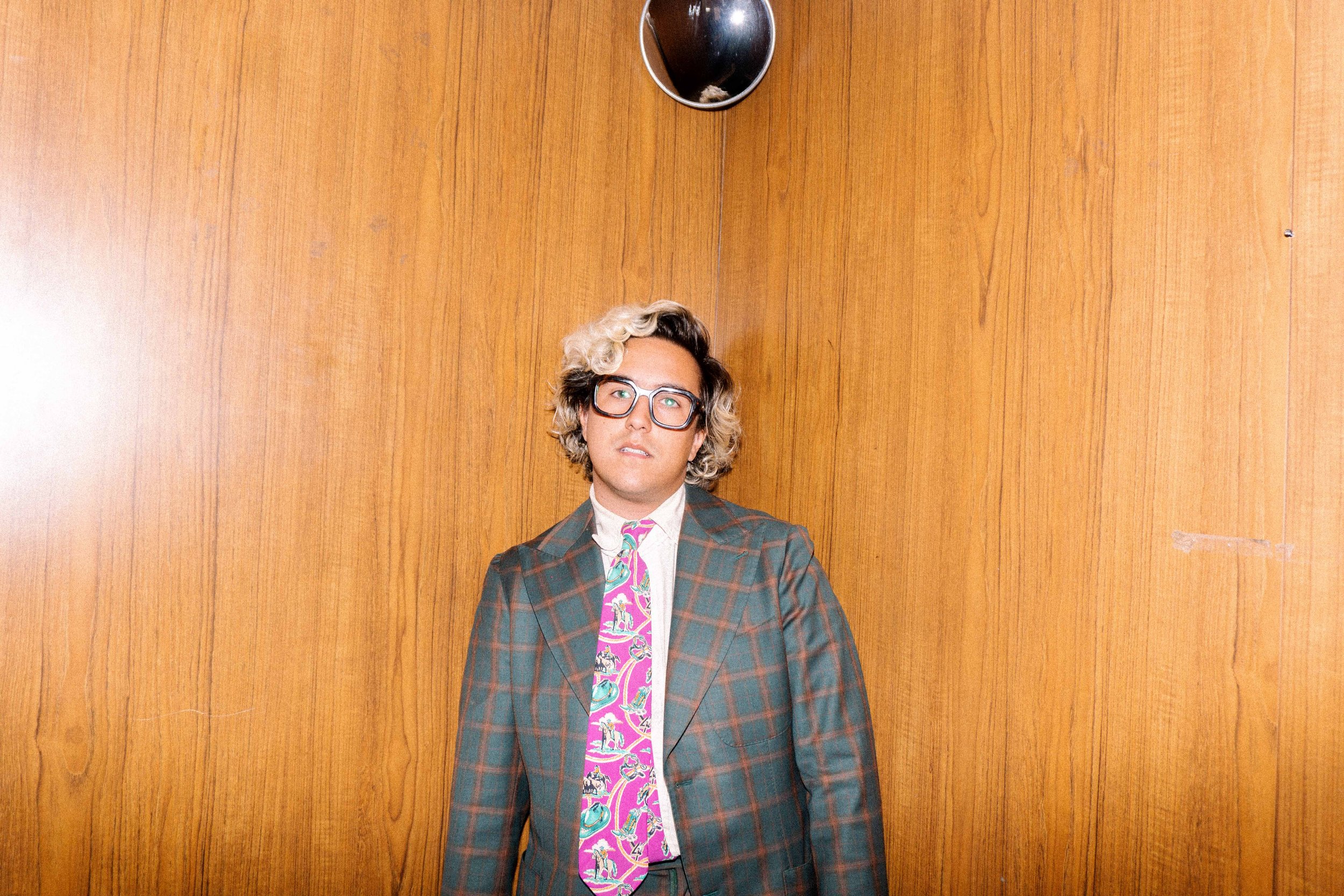 A young man with curly blond hair, glasses, wearing a colorful plaid suit and a pink tie with a whimsical pattern, standing against a wooden wall.