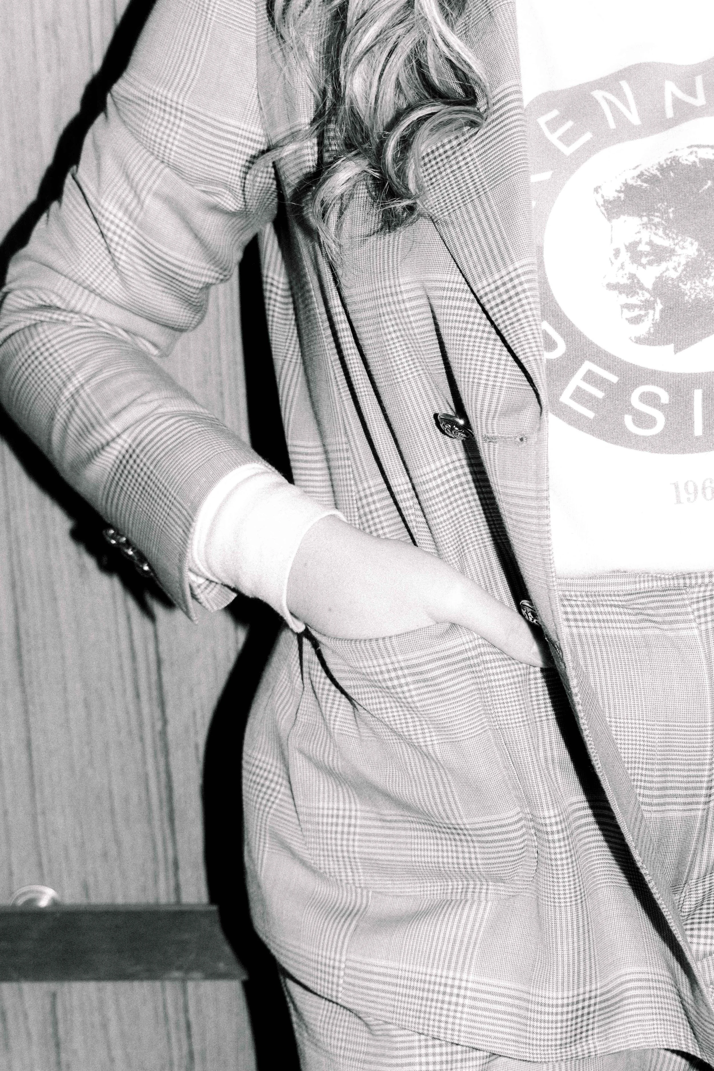 Close-up of a person wearing a checkered jacket, t-shirt, and carrying a bag, standing against a wooden background.