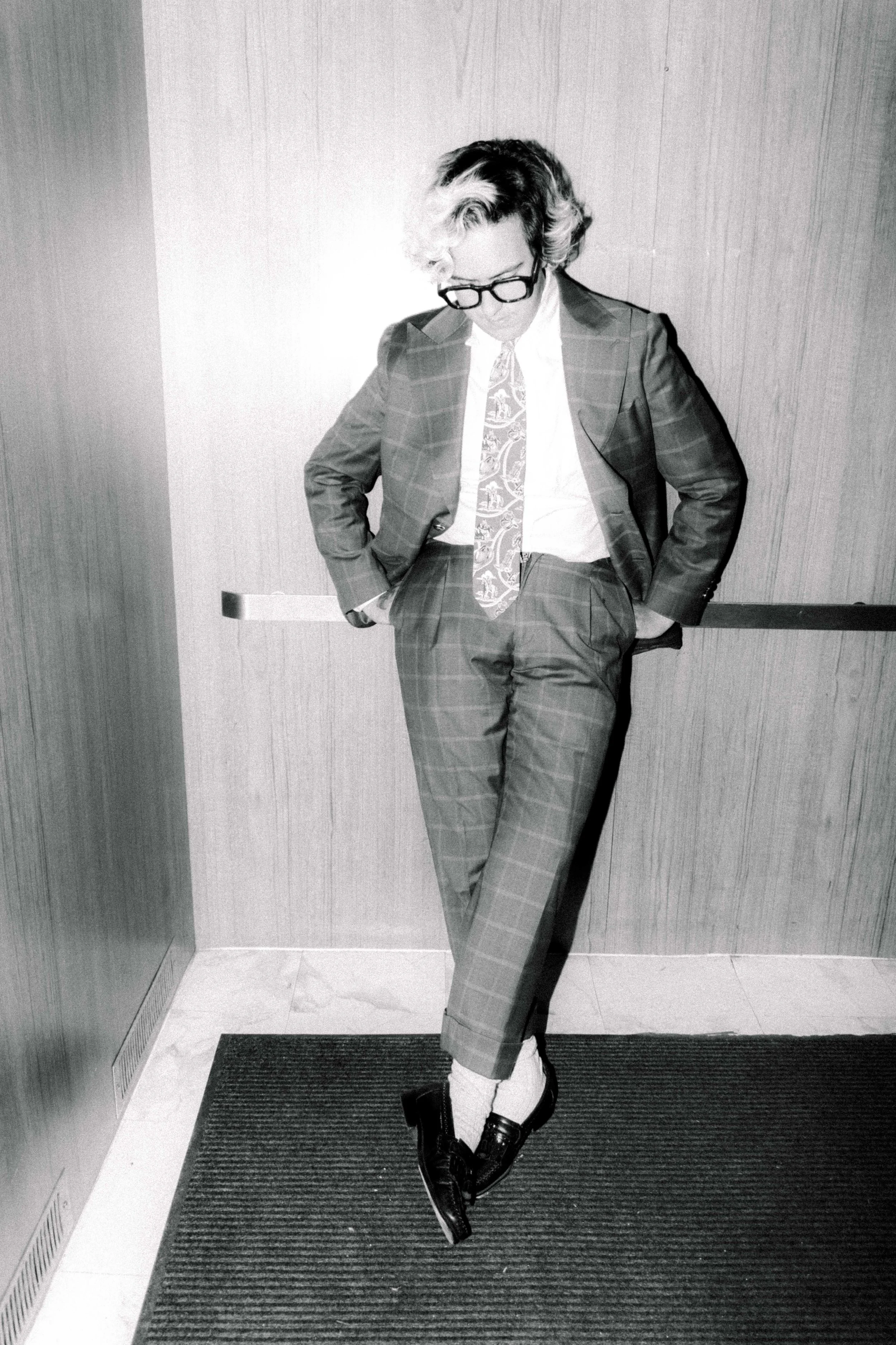 A person dressed in a checkered suit, white shirt, patterned tie, and glasses, standing with one foot crossed over the other and hands in pockets against a wood-paneled wall.