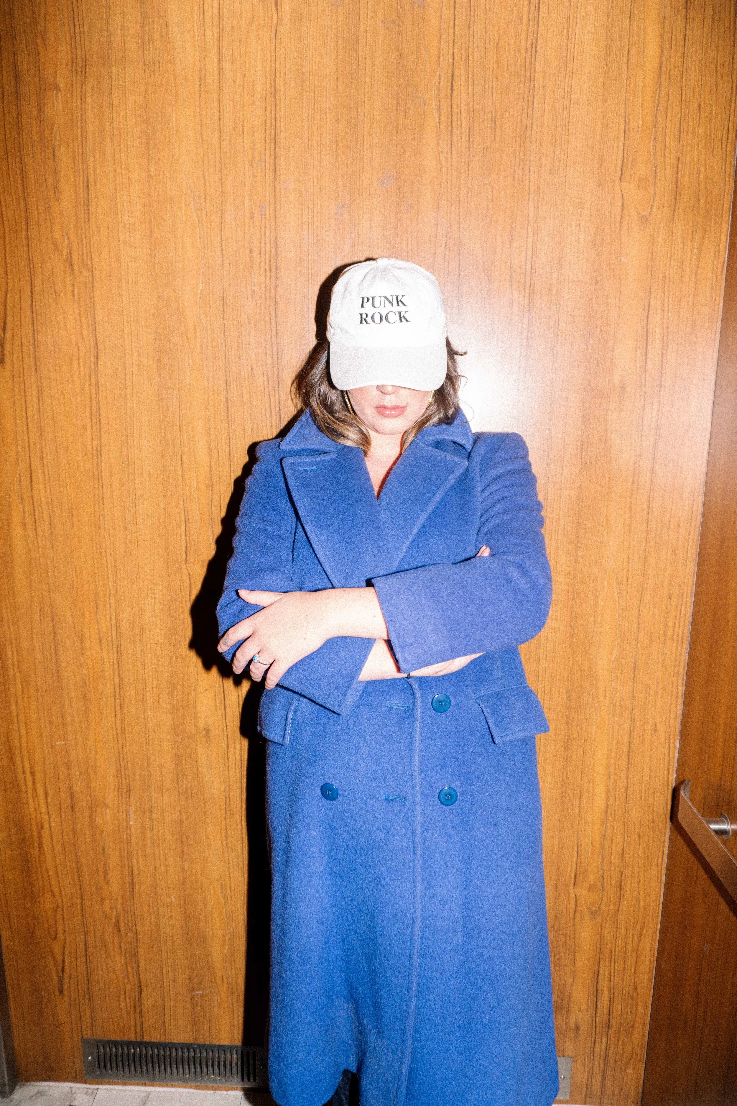 Woman wearing a blue coat and a beige cap with 'PUNK ROCK' written on it, standing against a wooden wall with crossed arms.