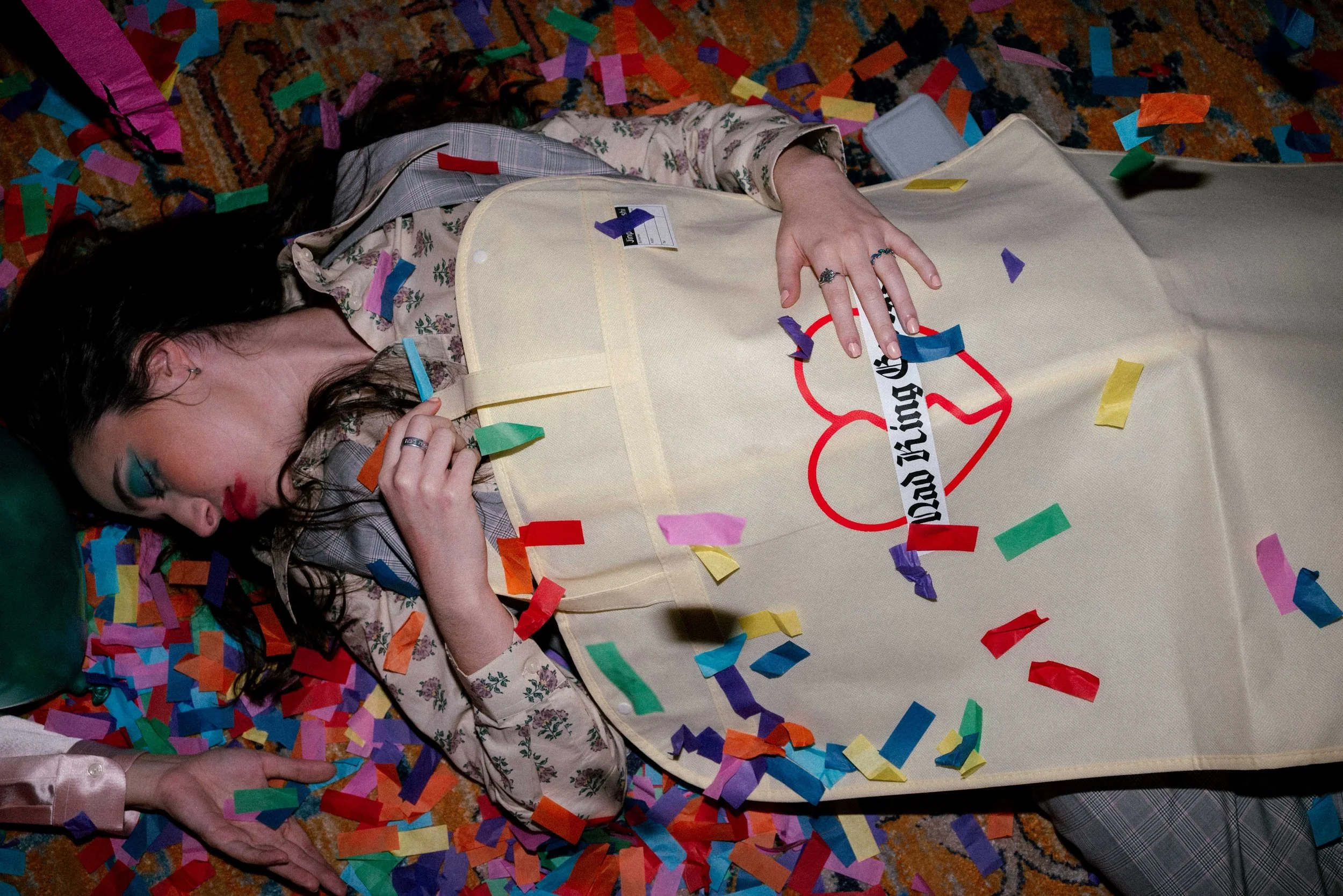 A woman lying on a colorful carpet surrounded by colorful confetti, with her shirt lifted to reveal a box with the text "Day Queen" and a red heart graphic. Her face shows makeup, and she has rings on her fingers.