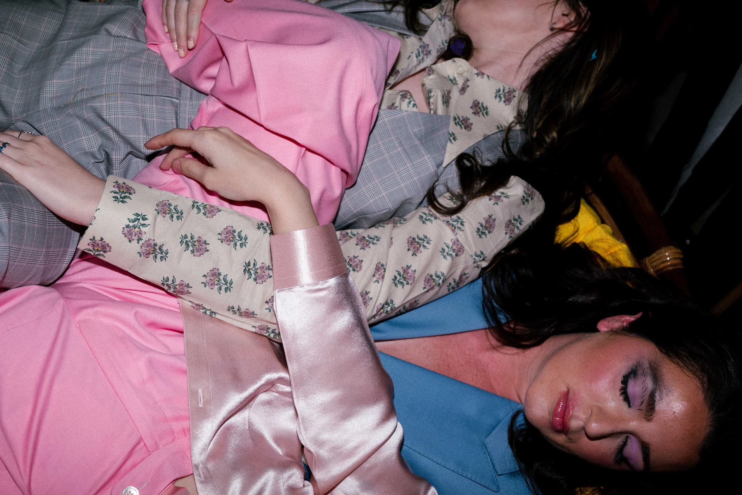 Two women lying close together in a dark room, dressed in colorful, patterned clothing, with one wearing pink satin and another in floral and plaid designs.