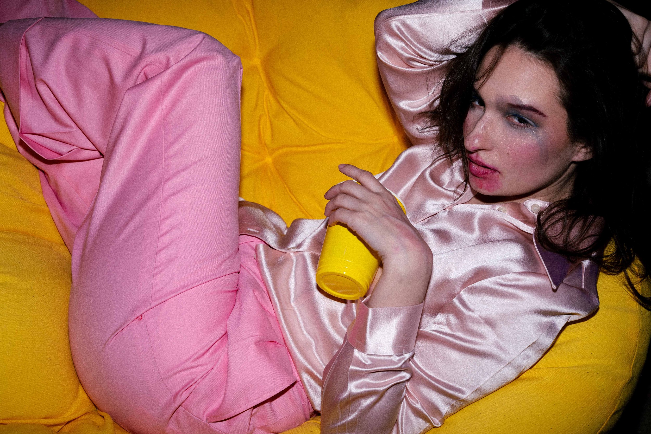 Woman lying on a yellow sofa, wearing pink satin pajamas, holding a yellow cup, with dark wavy hair and makeup.
