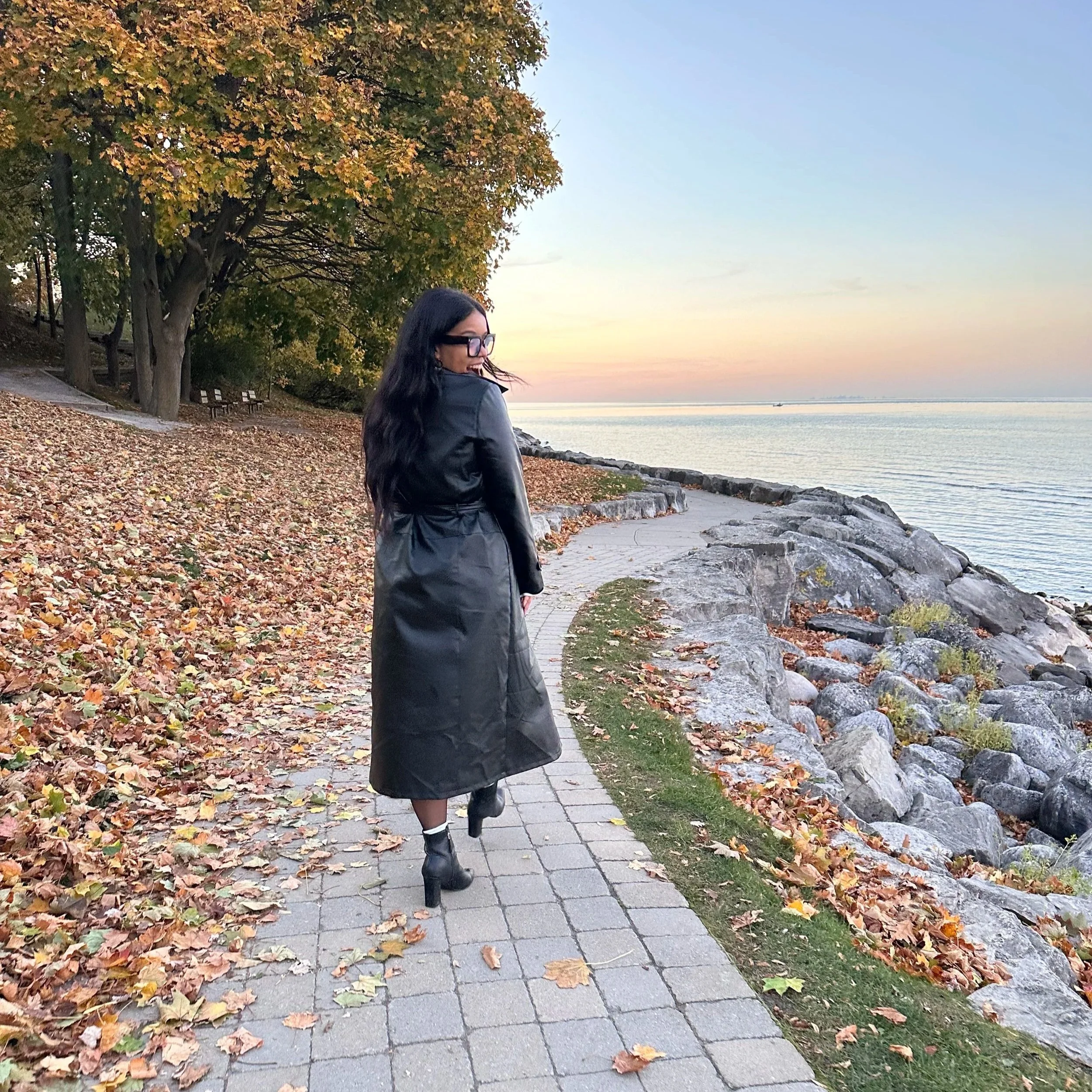 A woman with long dark hair, glasses, and black boots stands near a lake, looking at the water. She is wearing a black jacket, with fall leaves on the ground around her. A large tree with orange and green leaves is behind her, with benches nearby under the tree. The sky is clear with a hint of sunset colors.