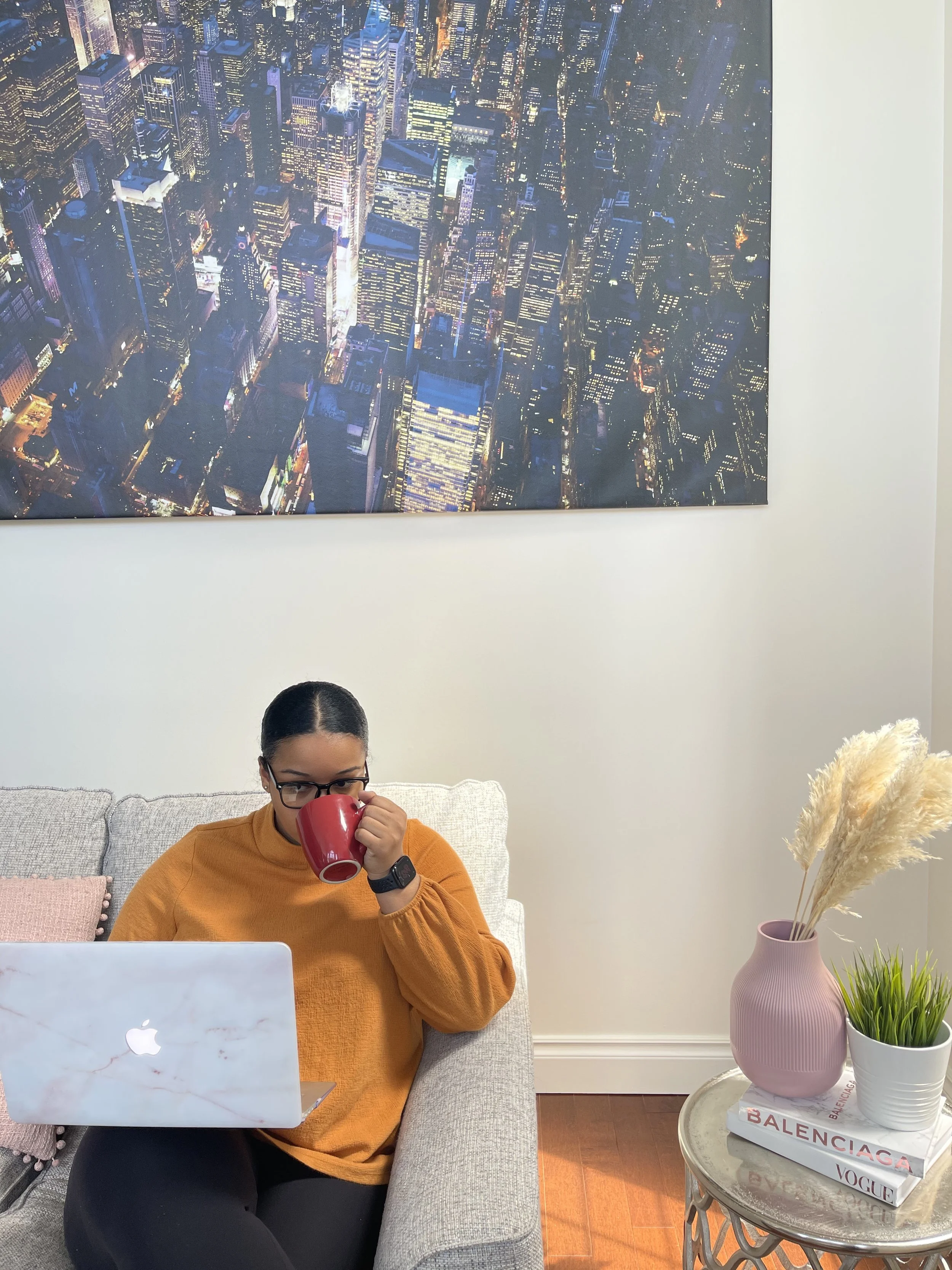 A woman with glasses and a smartwatch, wearing an orange sweater, sitting on a beige sofa, drinking from a red mug, with a MacBook laptop in front of her, a coffee table with books titled 'Balenciaga,' 'Vogue,' and 'Balenciaga,' and a pink vase with dried pampas grass and green plants beside her, under a cityscape photograph hanging on the wall.