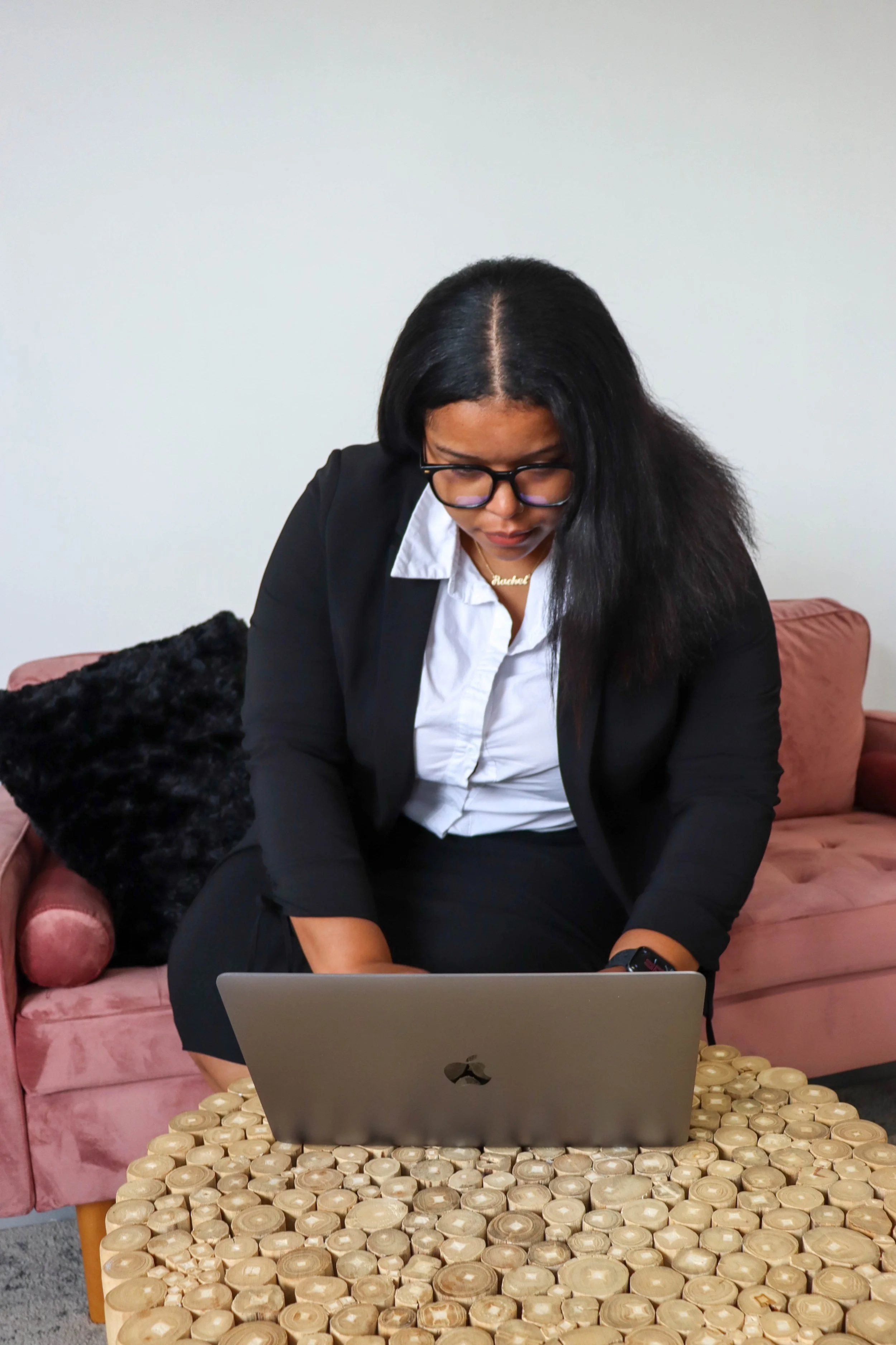 A woman with long black hair, glasses, and a suit, sitting on a pink velvet sofa with black pillows, leaning forward and working on a laptop. In front of her is a table covered with large round wood blocks, with a laptop resting on it.