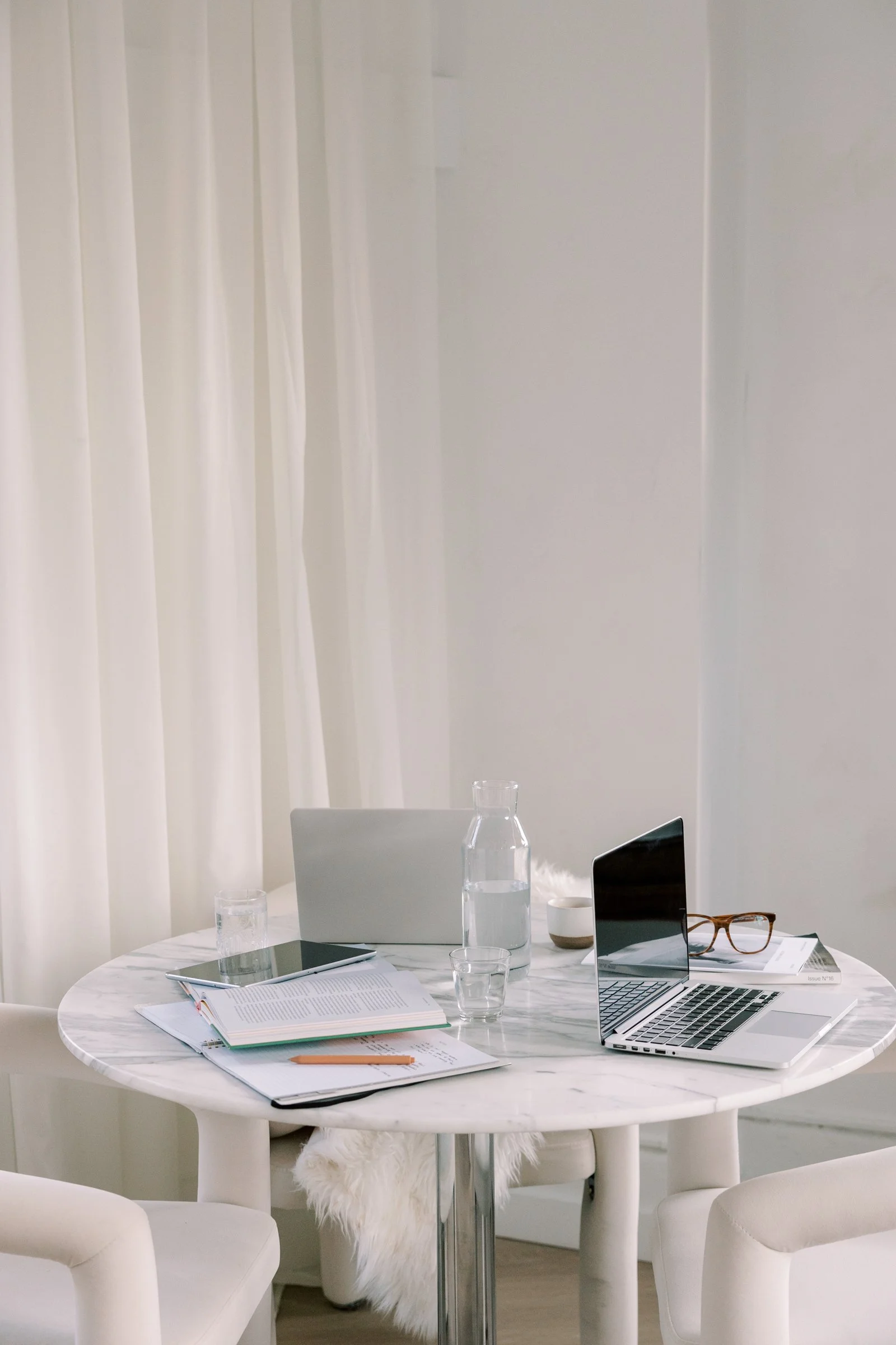 A round marble table with open notebooks, glasses, a laptop, a tablet, a pair of glasses, and a pencil. In the background, there is a white curtain and a white wall.
