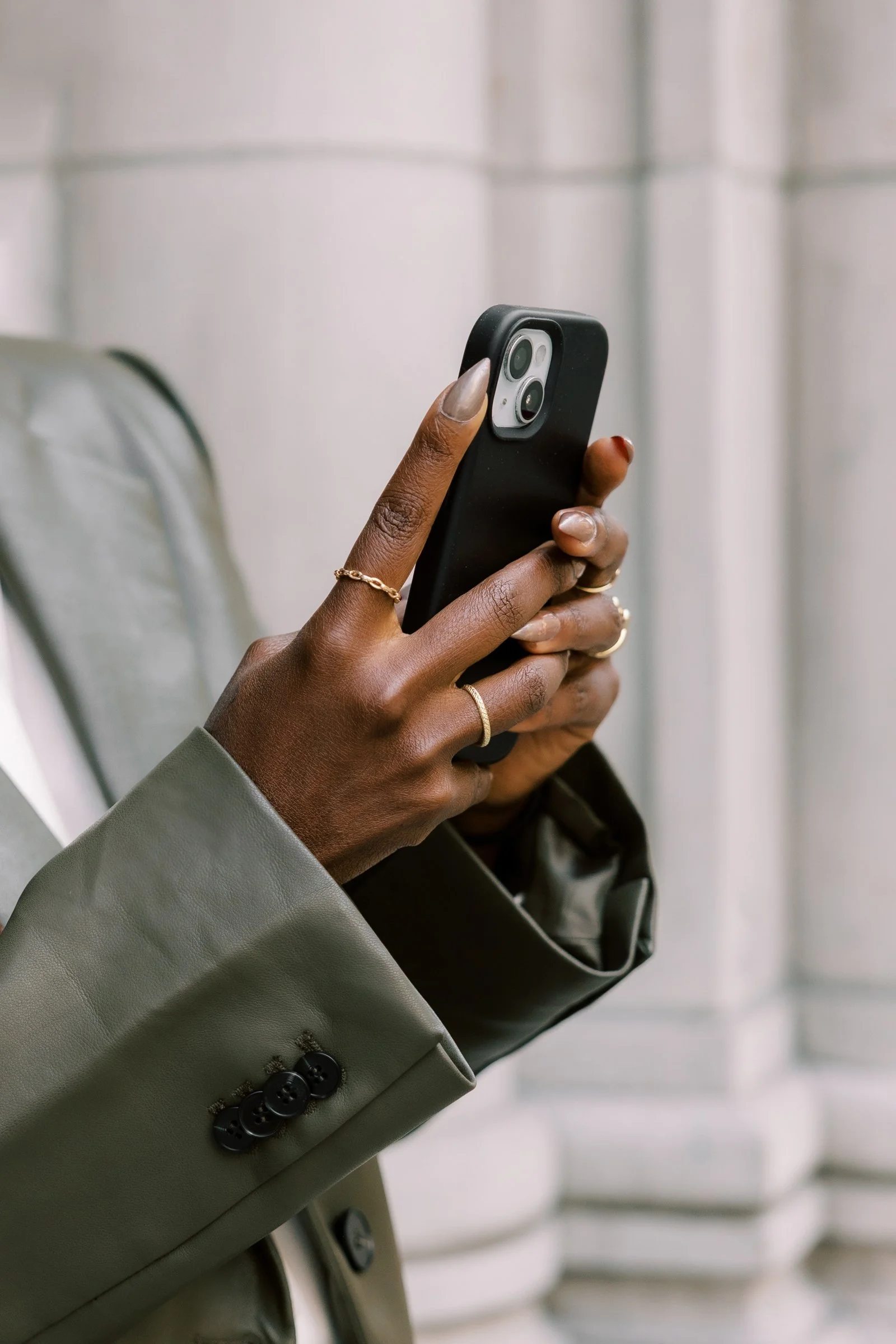 Person holding a black smartphone, wearing a gray blazer with button details, multiple rings on fingers, with a blurred background of white panels.