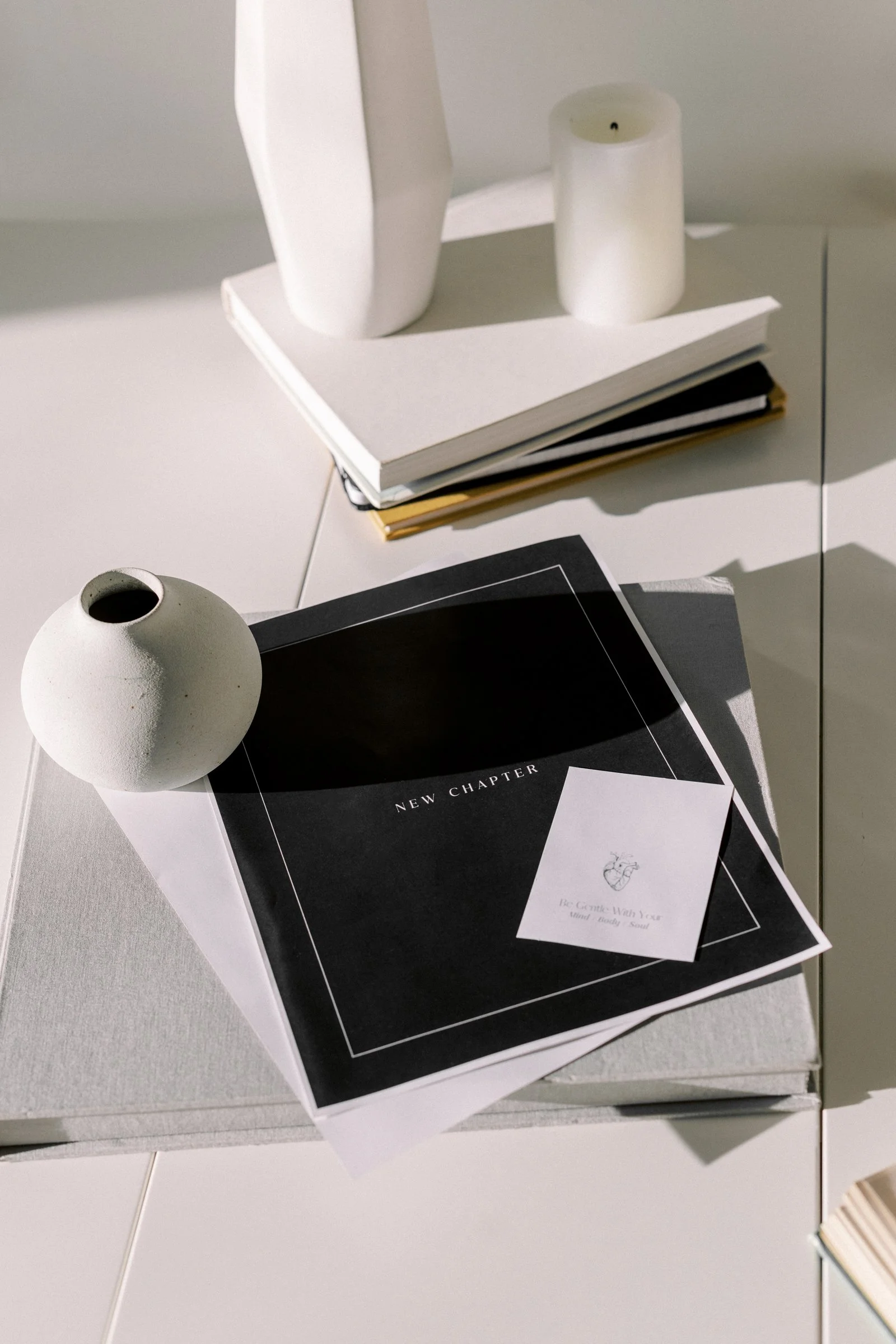 A white spherical vase with a round opening, a black and white magazine titled 'New Chapter,' a stack of papers or notebooks on top of a gray surface, a black and white booklet with text, and a partially visible open book in a beige or light-colored cover.