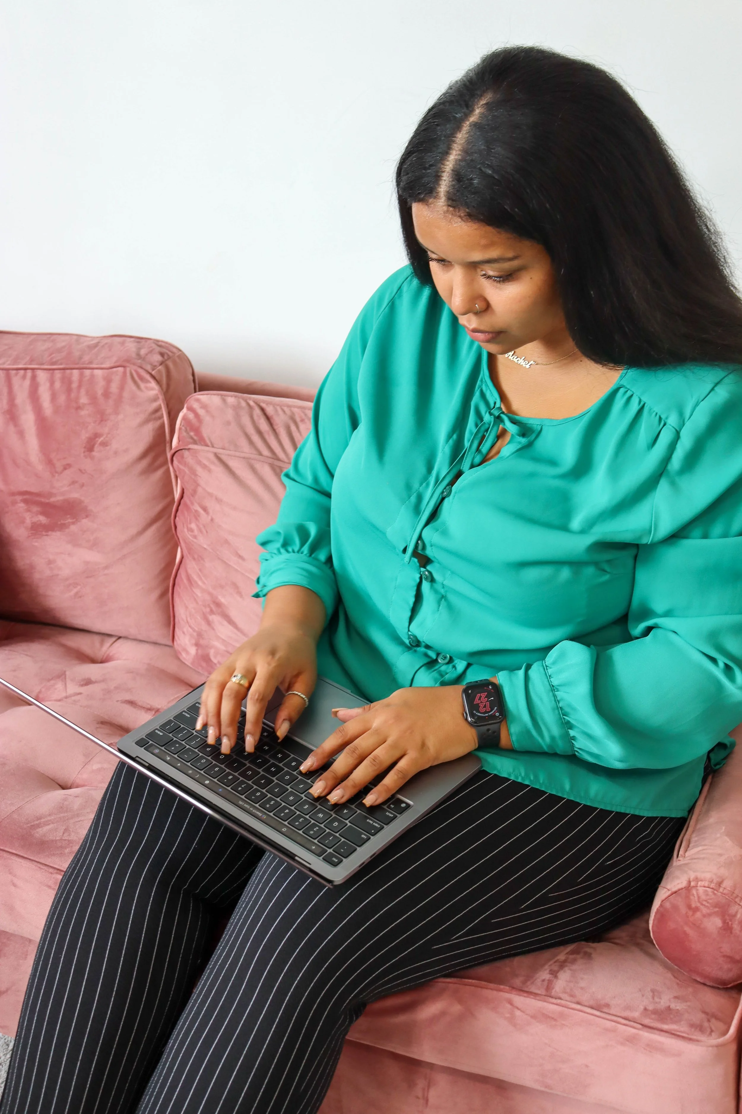 A woman with black hair, wearing a green blouse with a tie at the neck, black and white pinstripe pants, is sitting on a pink velvet couch. She is typing on a laptop on her lap, with a smartwatch on her left wrist and a nose ring.