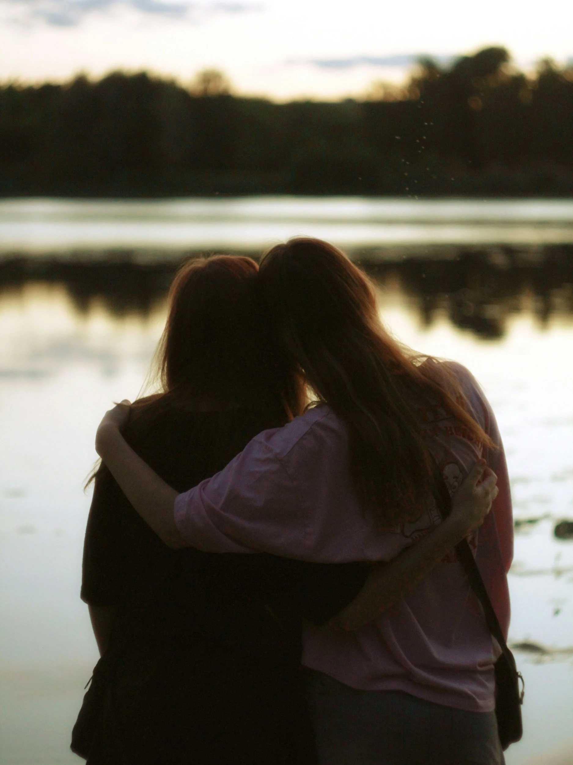 Two people embracing by a body of water, with trees and a sunset or sunrise sky in the background.