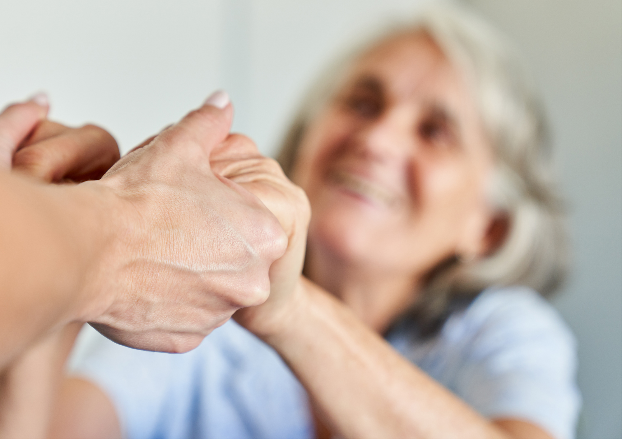 A smiling elderly woman with white hair holding a person's hand in a gesture of comfort or farewell, with a blurred background.