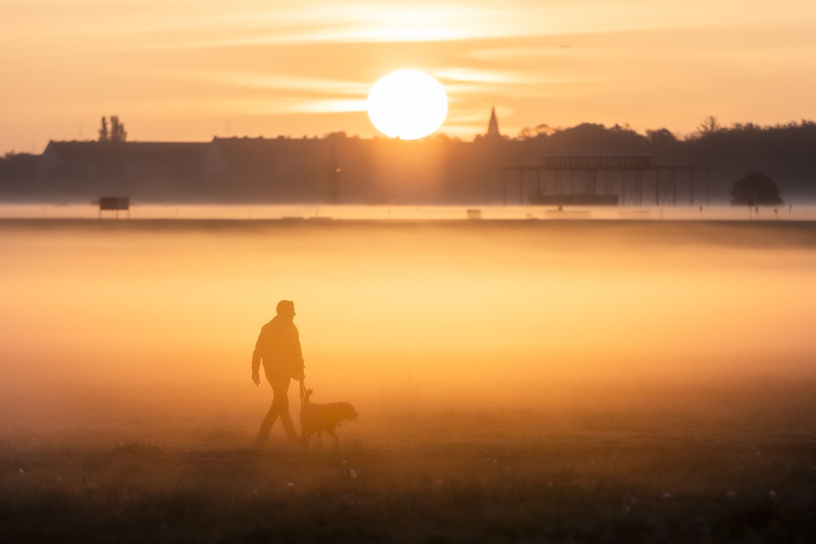 Ein Mann spaziert bei Sonnenaufgang und Nebel mit einem Hund auf dem Tempelhofer Feld in Berlin, 27.10.2022.