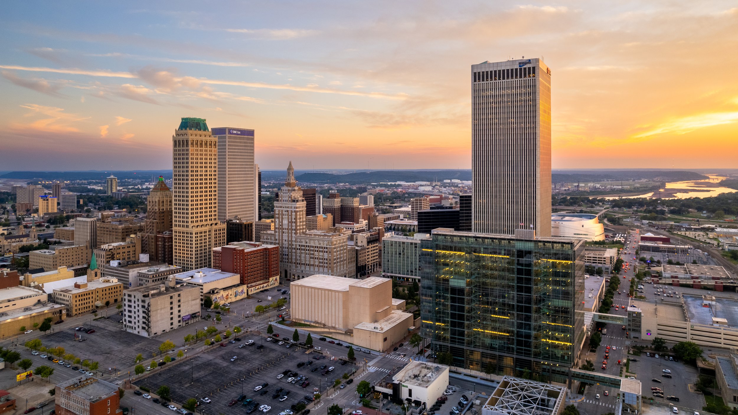 Aerial view of a cityscape during sunset with tall modern skyscrapers, shorter historic buildings, parking lots, and a river in the background.