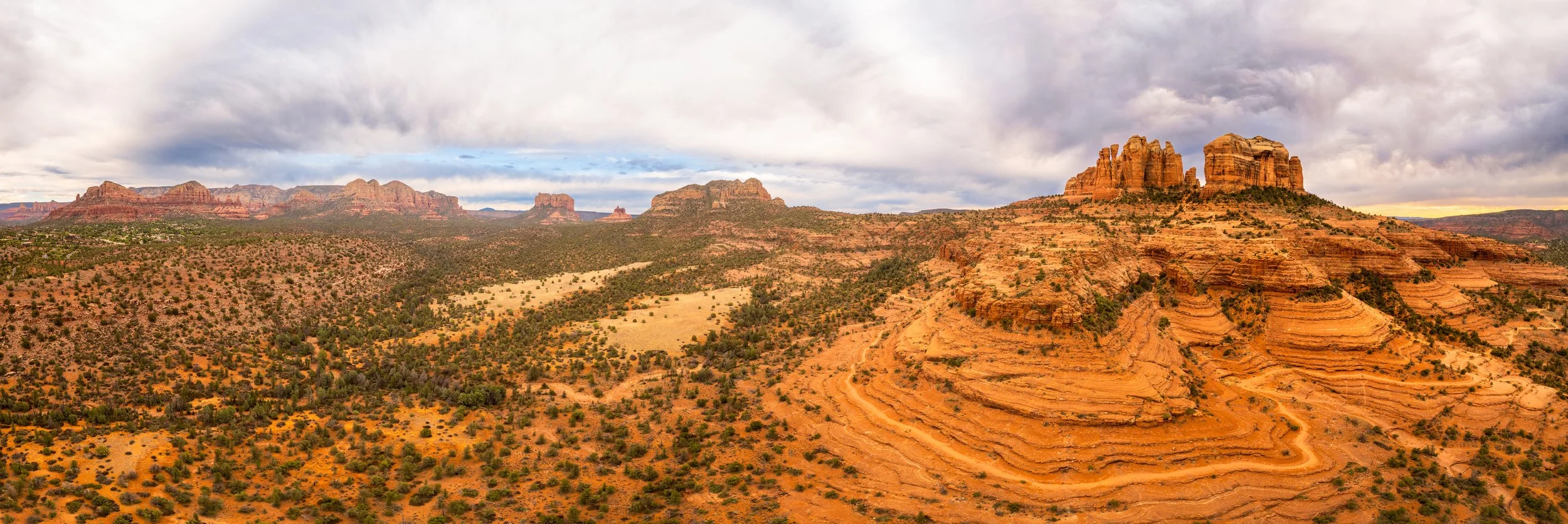 A panoramic view of Sedona, Arizona with red rock formations, including a prominent layered cliff on the right, under a cloudy sky.