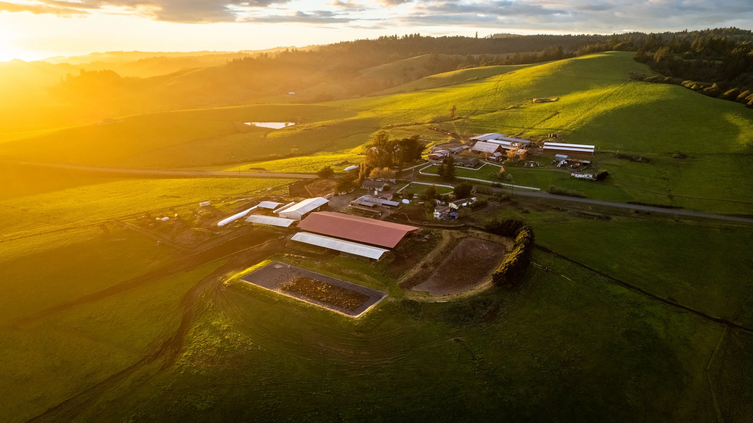 An aerial view of a farmstead with multiple buildings and green fields under a setting sun.
