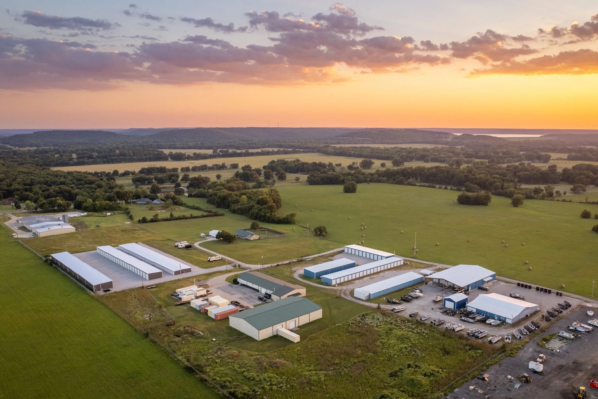 Aerial view of a farm at sunset with multiple blue and white barns, open fields with hay bales, and a distant horizon with a colorful sky.