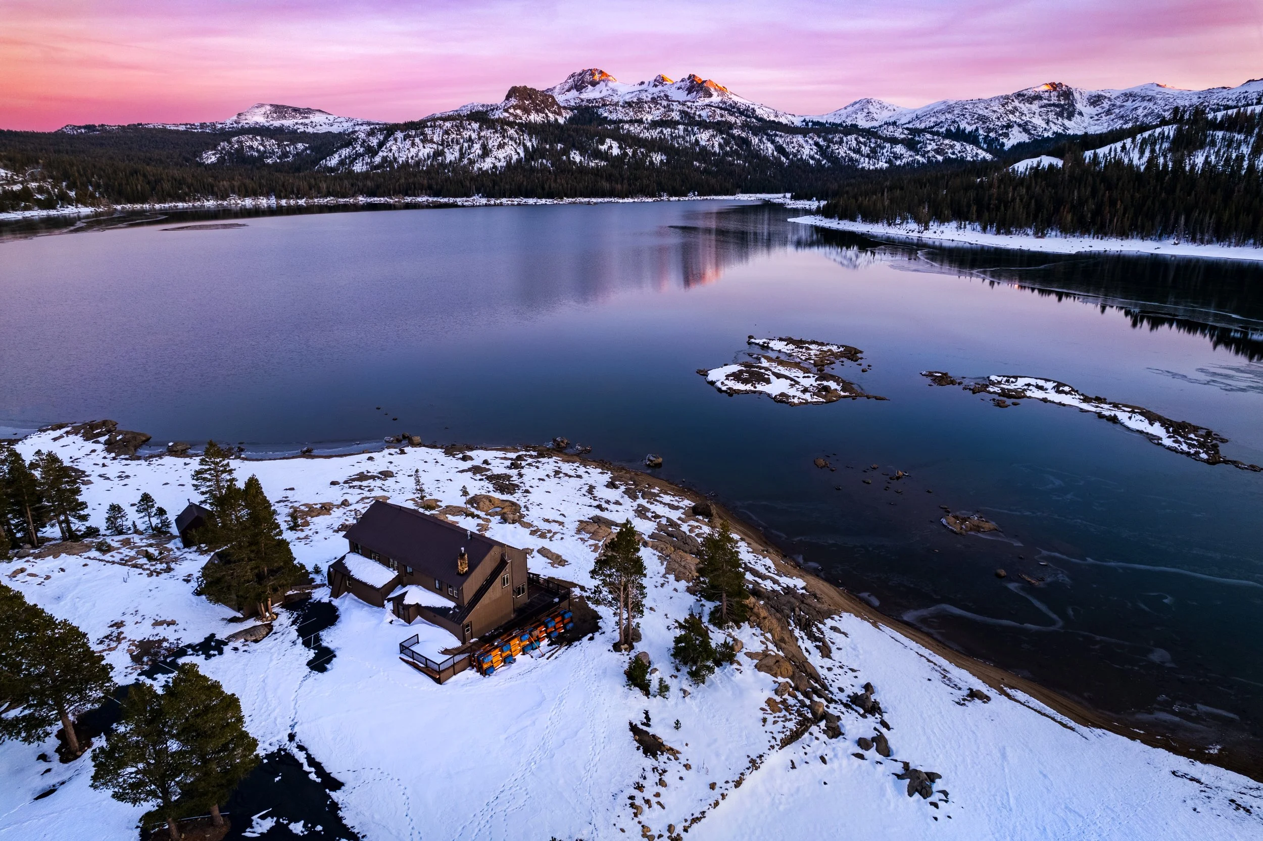 A snow-covered lakeside cabin surrounded by trees with a mountain range and a calm lake reflecting the sunset sky in the background.