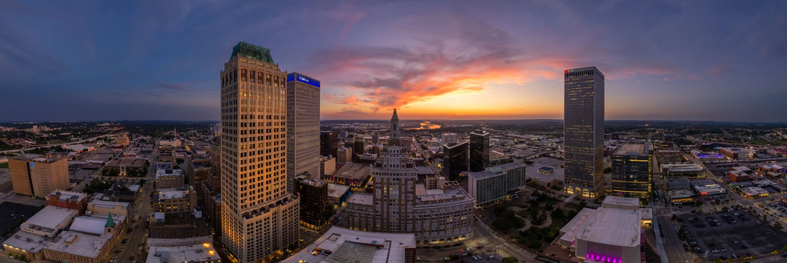 A city skyline at sunset with tall skyscrapers, a clock tower, and a mix of modern and historic buildings under a colorful sky.
