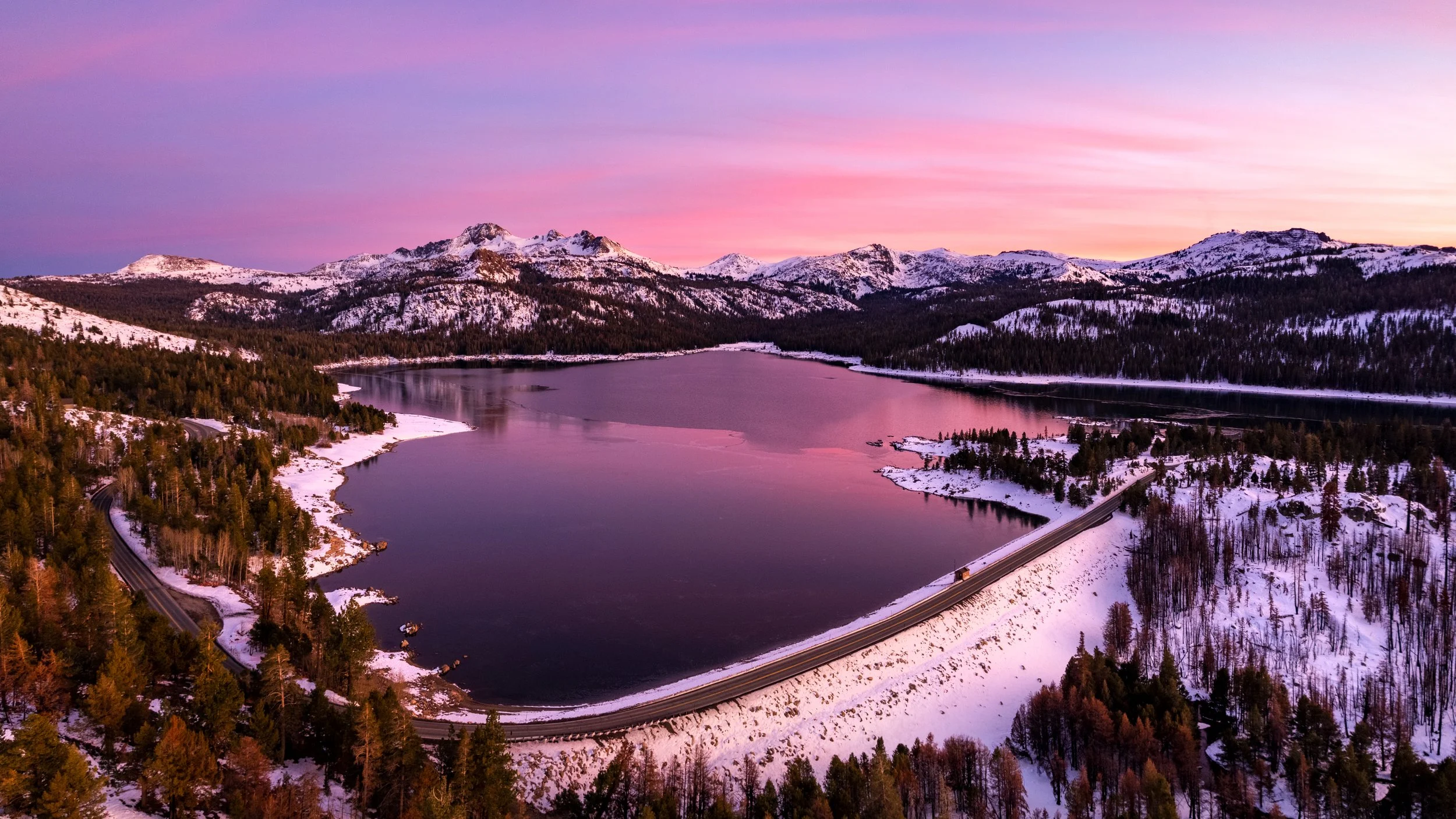 A scenic winter landscape featuring a large lake surrounded by snow-covered trees, mountains, and a sky with pink and purple hues at sunset.
